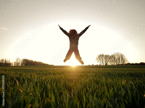 Joyful and happy girl is jumping with open arms on a green field at sunset with the sun in the background