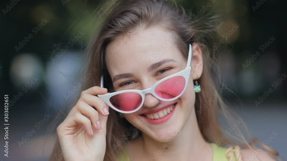 Portrait of beautiful girl wearing pink sunglasses smiling laughing to camera street outdoors. Happy young woman face close up Portrait of beautiful girl wearing pink sunglasses smiling laughing to camera street outdoors. Happy young woman face close up
