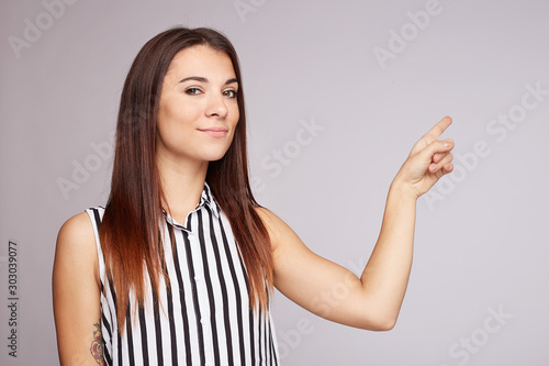 Pretty young brunette woman with positive expression, dressed casually, indicates with index finger at blank copy space for your promotional text or advertisement.  Adorable woman poses indoor alone.