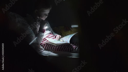 Girl resting in bed and reading aloud with the help of lantern. Little girl is reading a book while lying in her bed. photo