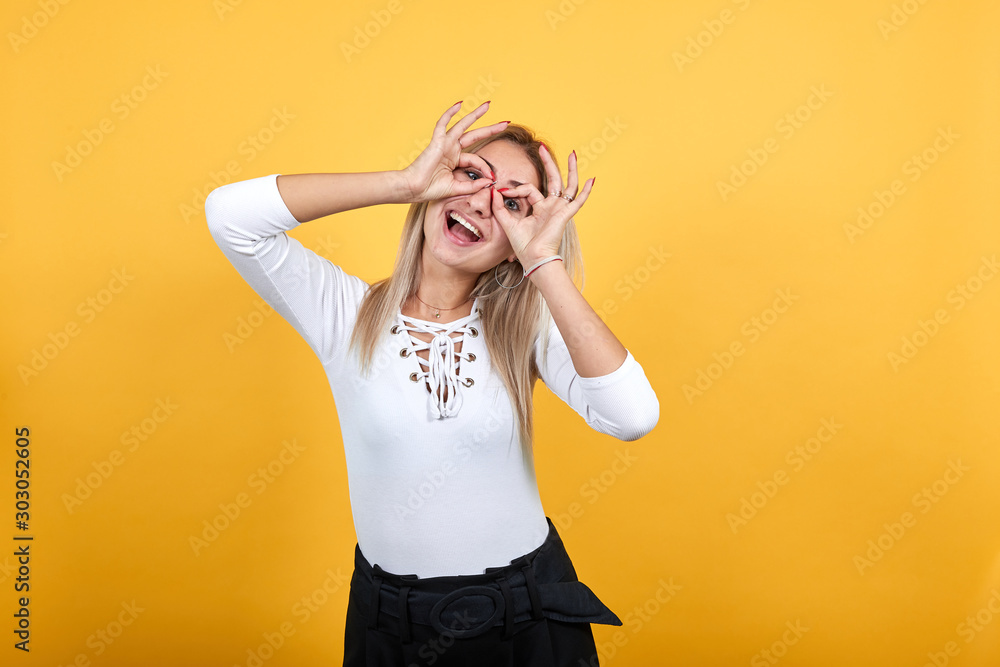 Young beautiful caucasian woman showing ok sign gesture near eyes loke glasses wearing white shirt on isolated orange background Young beautiful caucasian woman showing ok sign gesture near eyes loke glasses wearing white shirt on isolated orange background