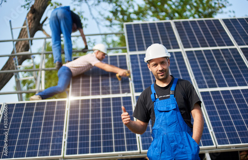 Portrait of smiling technician showing thumb up gesture standing in front of unfinished high exterior solar panel photo voltaic system, blue shiny surface with team of workers on high platform.