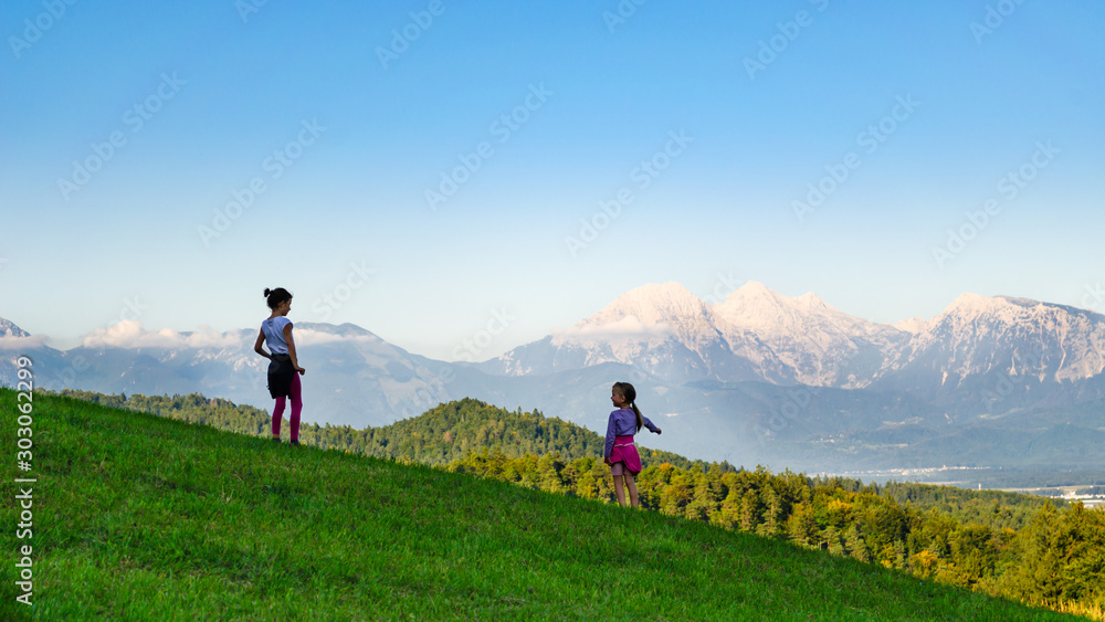 Obraz premium Rear view of two kid girls standing in a mountain meadow, younger one pointing to the valley below