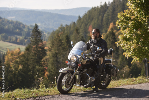 Handsome bearded biker in black leather jacket and sunglasses sitting on modern motorcycle on country roadside  on blurred background of foggy green hills covered with dense spruce forest.