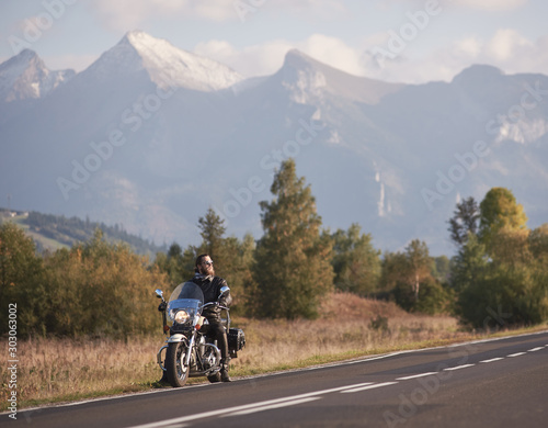 Young bearded biker in black leather jacket and sunglasses sitting on bike on country roadside on blurred background of green rural landscape  distant white sunny mountain peaks and bright sky.