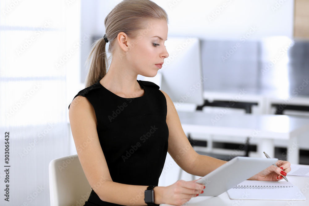 Business woman working with tablet computer while sitting at the desk in modern office. Secretary or female lawyer looks beautiful in black dress. Business people concept Business woman working with tablet computer while sitting at the desk in modern office. Secretary or female lawyer looks beautiful in black dress. Business people concept