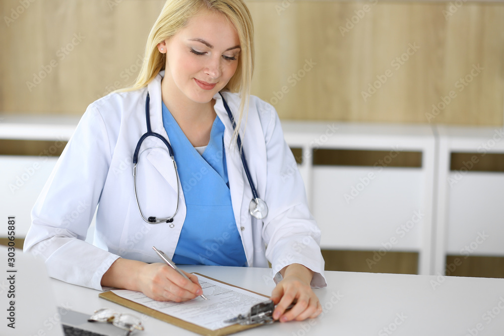 Doctor woman at work while sitting at the desk in hospital or clinic. Blonde cheerful physician filling up medical records form. Data and best service in medicine and healthcare Doctor woman at work while sitting at the desk in hospital or clinic. Blonde cheerful physician filling up medical records form. Data and best service in medicine and healthcare