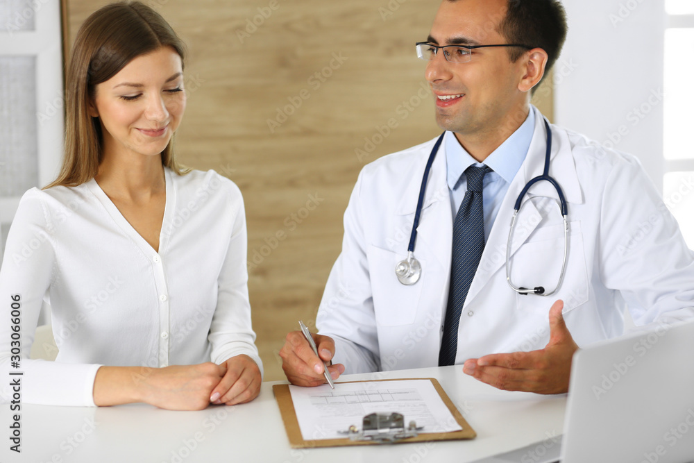 Doctor and patient discussing the results of a physical examination while sitting at a desk in a clinic. A male doctor using a clipboard to fill out a medical history of a young woman's medication Doctor and patient discussing the results of a physical examination while sitting at a desk in a clinic. A male doctor using a clipboard to fill out a medical history of a young woman's medication