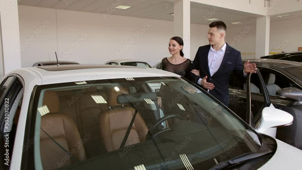Young couple watching a new white car in the showroom of the dealership. Happy young couple choose a new modern car in the showroom. Young couple watching a new white car in the showroom of the dealership. Happy young couple choose a new modern car in the showroom.