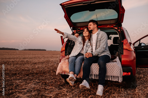 Young Happy Couple Dressed Alike in White Shirt and Jeans Sitting at Their New Car Trunk, Beautiful Sunset on the Field, Vacation and Travel Concept