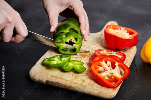 Pepper slicing. Hands, knife and red, green, yellow bell peppers, wooden cutting board on stone table. Sliced sweet peppers on black background. Vegetable ingredient, cooking diet salad, healthy food