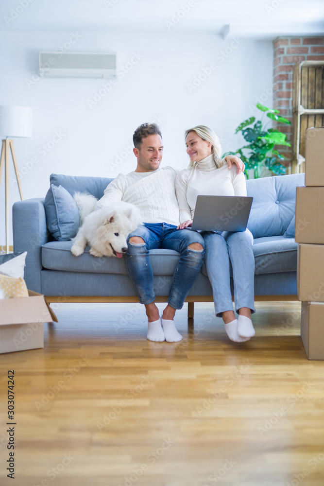Young beautiful couple with dog sitting on the sofa using laptop at new home around cardboard boxes Young beautiful couple with dog sitting on the sofa using laptop at new home around cardboard boxes