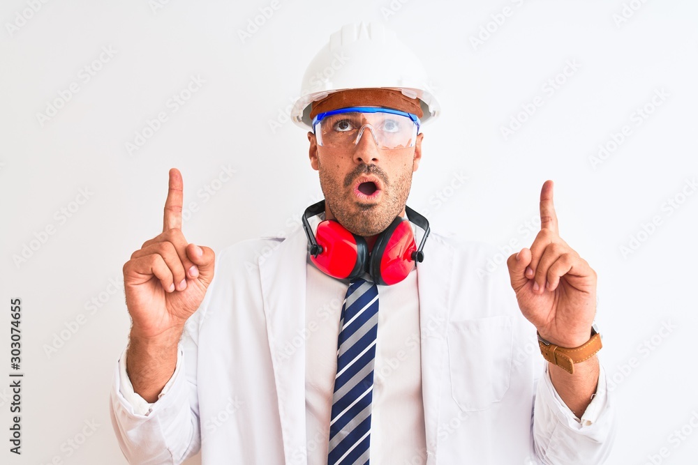 Young chemist man wearing security helmet and headphones over isolated background amazed and surprised looking up and pointing with fingers and raised arms.