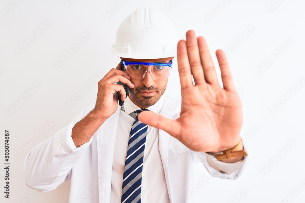Young scientist man wearing safety helmet and calling using smartphone over isolated background with open hand doing stop sign with serious and confident expression, defense gesture Young scientist man wearing safety helmet and calling using smartphone over isolated background with open hand doing stop sign with serious and confident expression, defense gesture