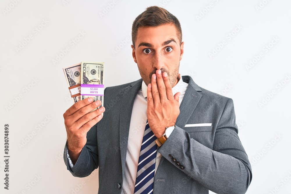 Young handsome business man holding bunch of dollars banknotes over isolated background cover mouth with hand shocked with shame for mistake, expression of fear, scared in silence, secret concept Young handsome business man holding bunch of dollars banknotes over isolated background cover mouth with hand shocked with shame for mistake, expression of fear, scared in silence, secret concept