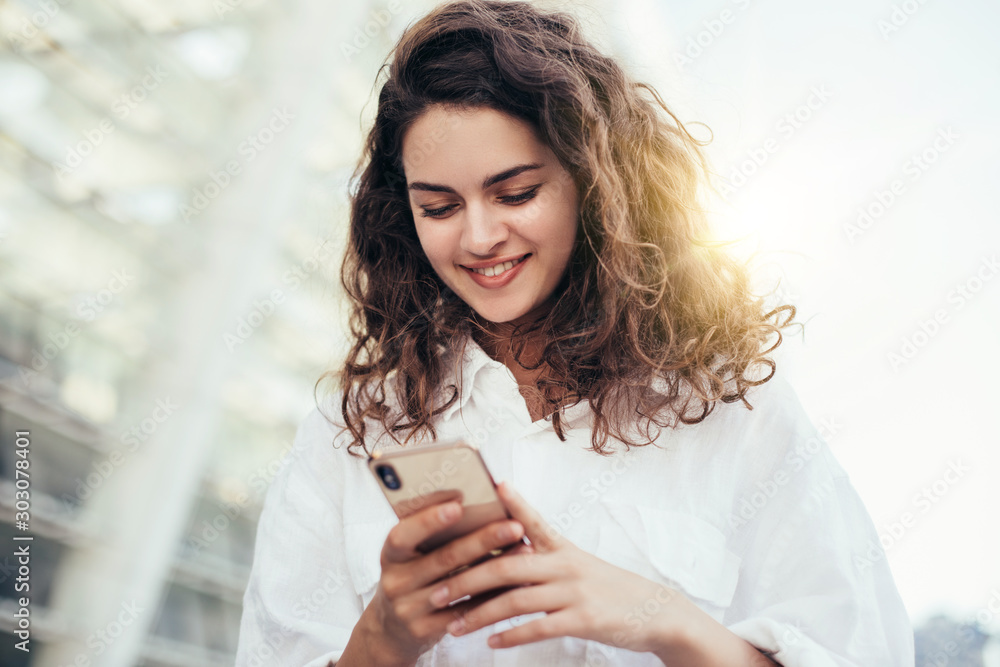 Positive attractive young brunette woman stand outside and look at phone in hands. White blouse and curly hair. Buildings behind. Sun shines and beautiful daylight. Positive attractive young brunette woman stand outside and look at phone in hands. White blouse and curly hair. Buildings behind. Sun shines and beautiful daylight.