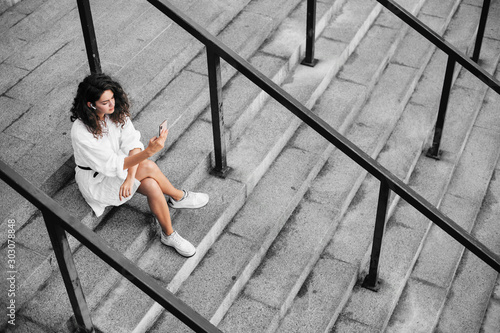 Calm peaceful and thoughtful young woman sit on grey steps and look on phone. Taking pictures alone. Wear white dress and shoes.