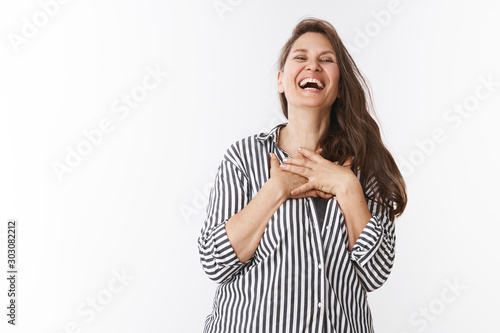 Beautiful middle aged woman in striped trendy blouse chuckling, laughing out loud holding hands on chest from amazement shaking from laugh and joy bending head backwards amused over white background