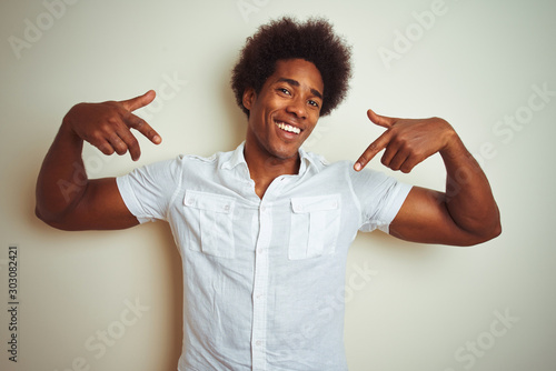 African american man with afro hair wearing shirt standing over isolated white background looking confident with smile on face, pointing oneself with fingers proud and happy.