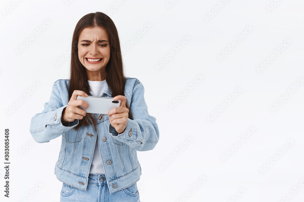 Intense pissed and angry pretty woman in denim jacket, playing mobile game, holding smartphone horizontally, grimacing as having intense battle online, standing white background pressured Intense pissed and angry pretty woman in denim jacket, playing mobile game, holding smartphone horizontally, grimacing as having intense battle online, standing white background pressured