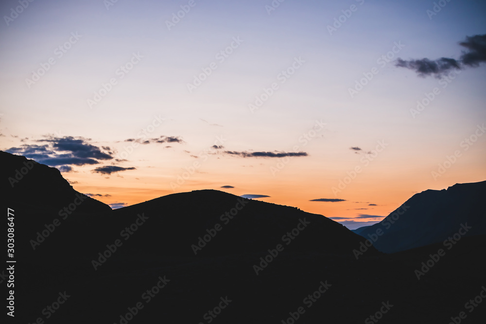 Atmospheric minimalist alpine landscape to big mountain silhouettes in sunset. Giant darker mountains under dawn sky with clouds. Vivid orange sundown. Wonderful highland scenery to multicolor dawn.