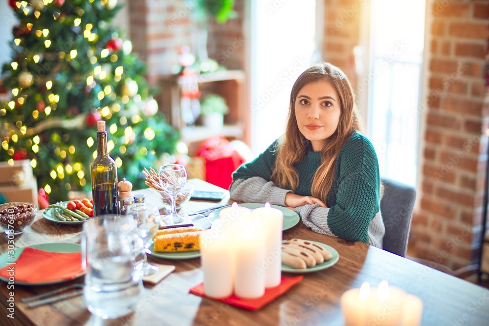Young beautiful woman sitting eating food around christmas tree at home with serious expression on face. Simple and natural looking at the camera.