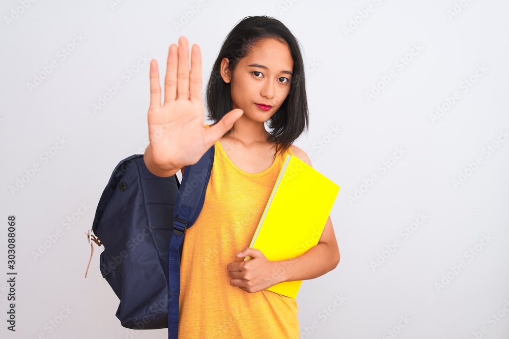 Young chinese student woman wearing backpack holding book over isolated white background with open hand doing stop sign with serious and confident expression, defense gesture Young chinese student woman wearing backpack holding book over isolated white background with open hand doing stop sign with serious and confident expression, defense gesture