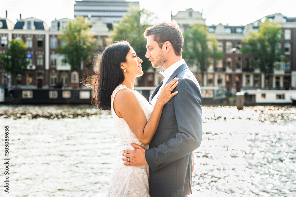 A happy young couple look at each other on their wedding day, standing in front of an Amsterdam canal