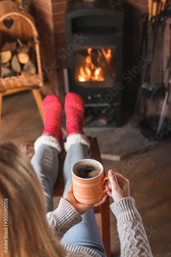 Woman is drinking hot coffee in front of fireplace and warming her feet with wool socks