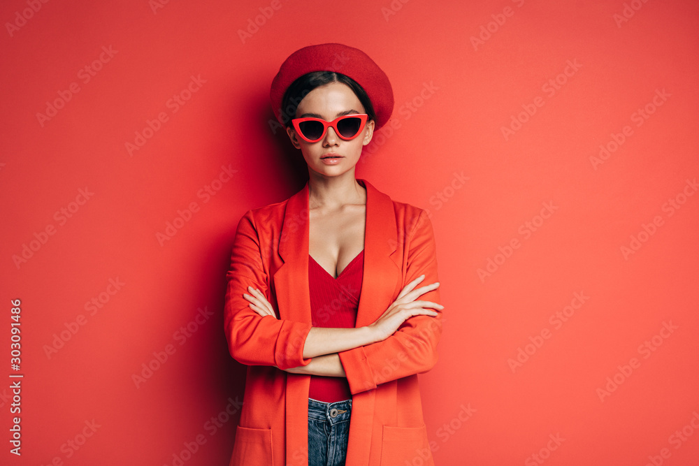 Serious concentrated model in xunglasses and red clothes posing on camera. Hands crossed. Look straight. Wear lacket and beret. Stylish modern model. Isolated over red baackground. Serious concentrated model in xunglasses and red clothes posing on camera. Hands crossed. Look straight. Wear lacket and beret. Stylish modern model. Isolated over red baackground.