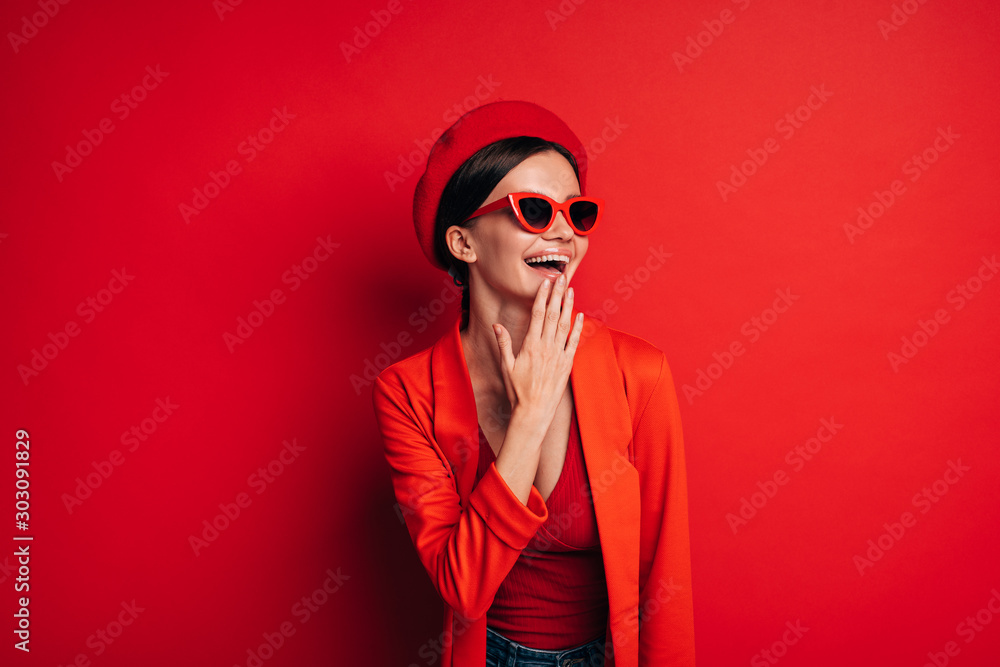 Beautiful young woman laughing. Try to cover mouth with hand. Stand alone in red clothes and beret. Look through sunglasses. Emotional cheerful model. Isolated over red background. Beautiful young woman laughing. Try to cover mouth with hand. Stand alone in red clothes and beret. Look through sunglasses. Emotional cheerful model. Isolated over red background.