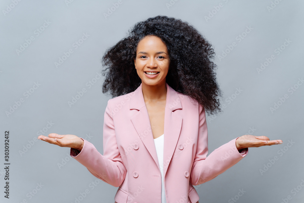 Isolated shot of cheerful female business worker with Afro hairstyle, wears elegant formal violet jacket, raises both palms, presents some product, smiles pleasantly, isolated over grey background. - obrazy, fototapety, plakaty 