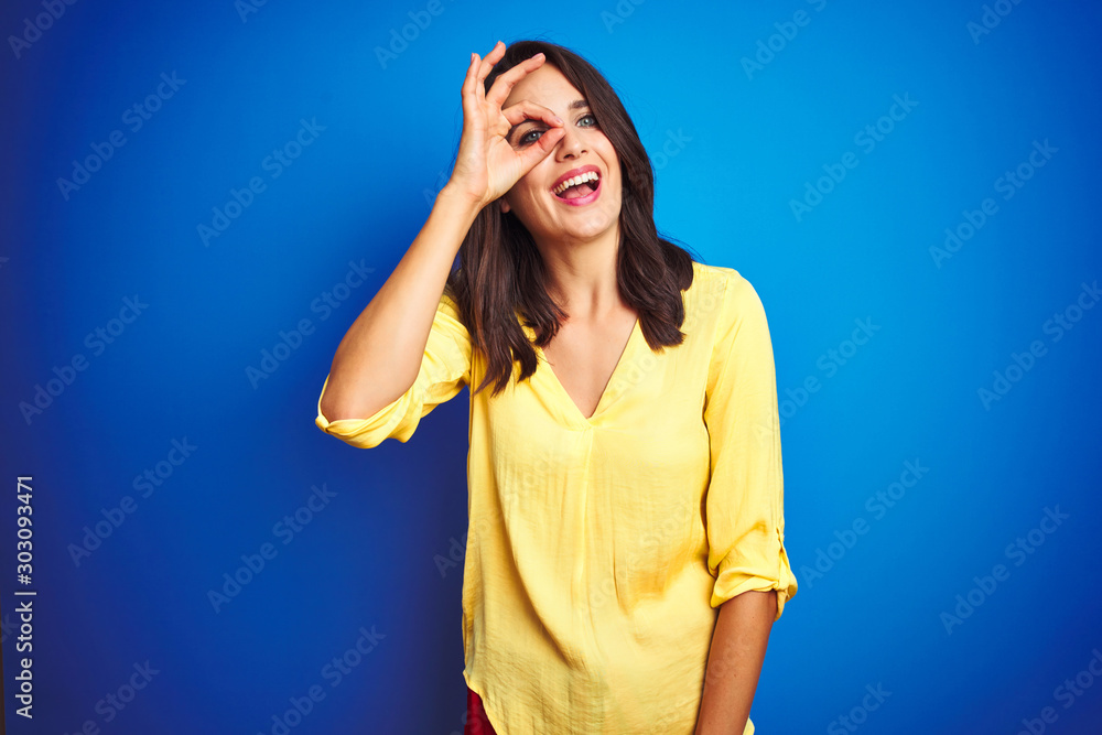 Young beautiful woman wearing yellow t-shirt standing over blue isolated background doing ok gesture with hand smiling, eye looking through fingers with happy face. Young beautiful woman wearing yellow t-shirt standing over blue isolated background doing ok gesture with hand smiling, eye looking through fingers with happy face.