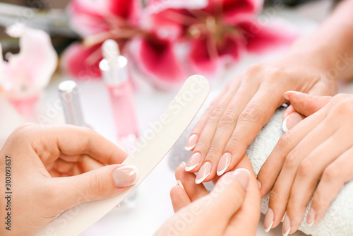 Woman beautician using a nail file. Professional and beautiful hands with nails care manicure applying in luxury salon. Pink red flowers background.