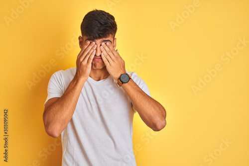Young indian man wearing white t-shirt standing over isolated yellow background rubbing eyes for fatigue and headache, sleepy and tired expression. Vision problem