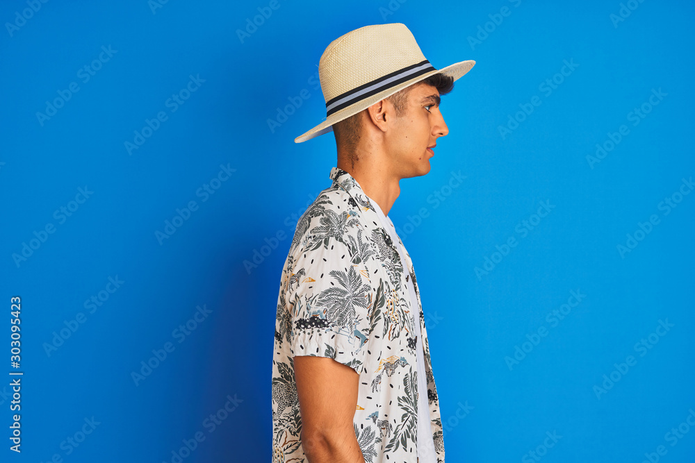 Indian man on vacation wearing hawiaian shirt summer hat over isolated blue background looking to side, relax profile pose with natural face with confident smile.