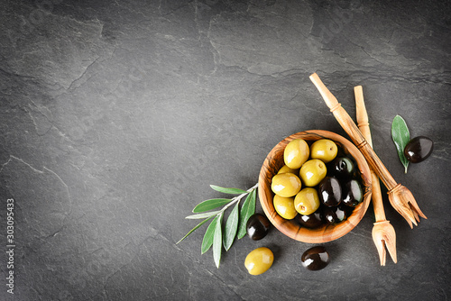 Fresh olives in wooden bowl on dark stone table. Olive branchs or leaves on black background.