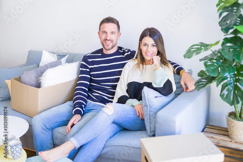 Young beautiful couple sitting on the sofa drinking cup of coffee at new home around cardboard boxes