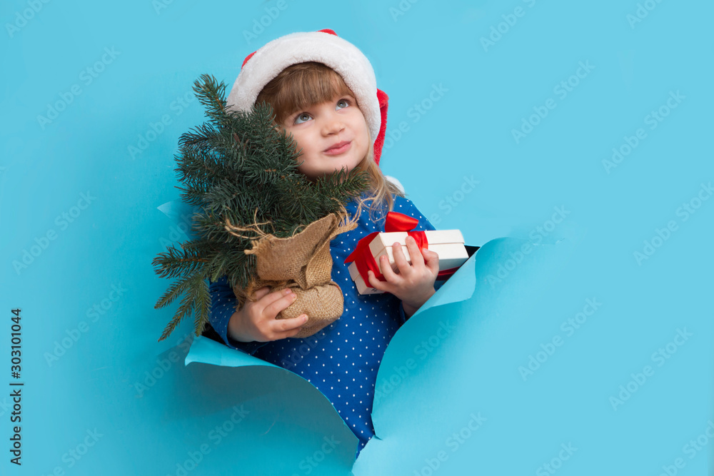 Obraz premium Happy smiling kid in Santa red hat with Christmas tree and gift poses through torn paper hole. Effect of torn paper. Christmas sales.