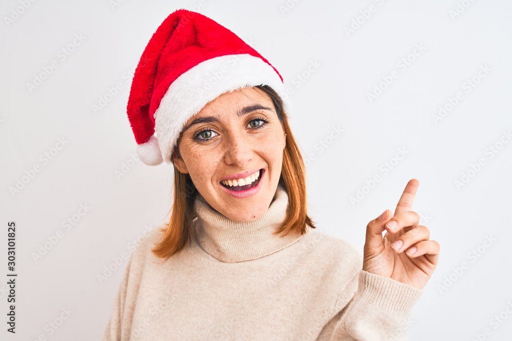 Beautiful redhead woman wearing christmas hat over isolated background very happy pointing with hand and finger to the side Beautiful redhead woman wearing christmas hat over isolated background very happy pointing with hand and finger to the side