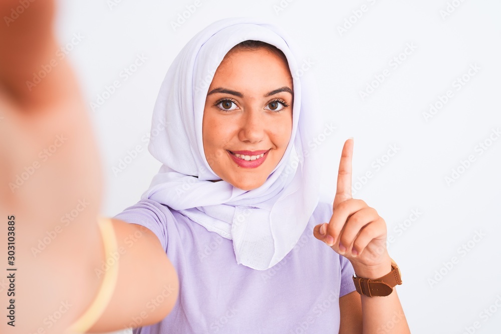 Beautiful arabian girl wearing hijab make selfie by camera over isolated white background surprised with an idea or question pointing finger with happy face, number one Beautiful arabian girl wearing hijab make selfie by camera over isolated white background surprised with an idea or question pointing finger with happy face, number one