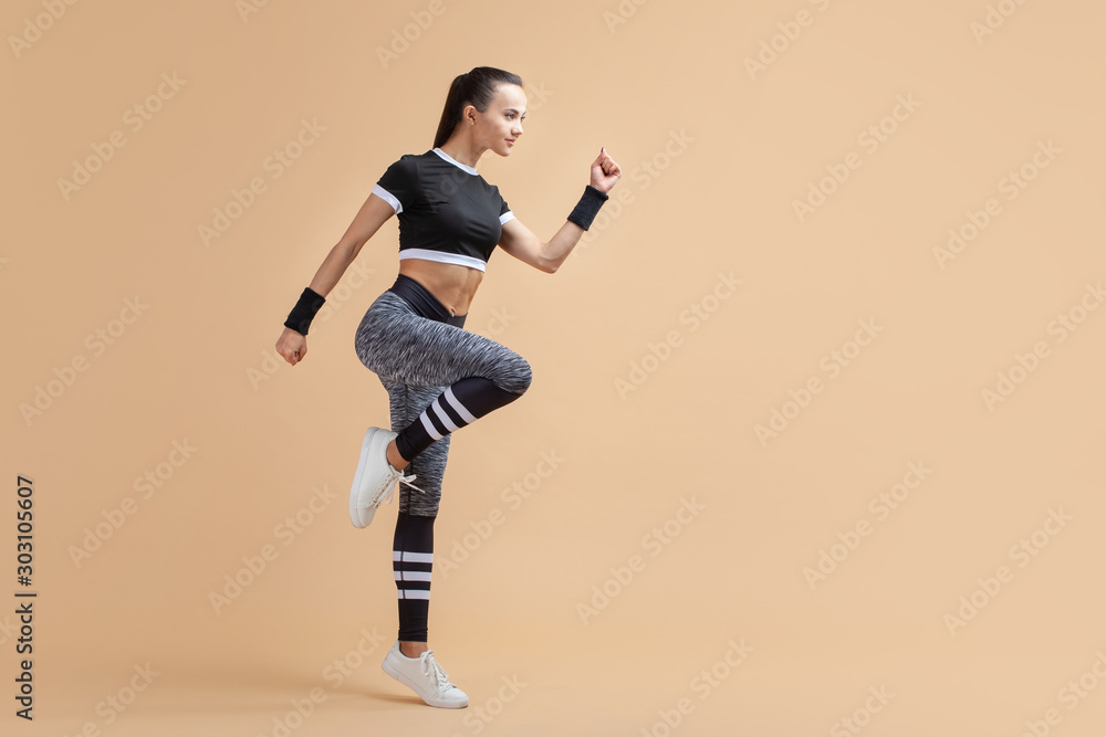 A young attractive brunette girl runs with wireless headphones raising her knees high on a peach background, cardio.