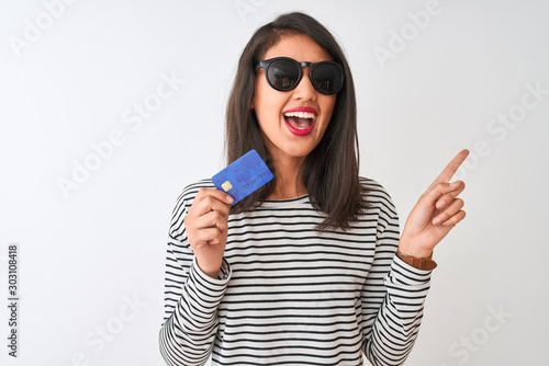 Young chinese woman wearing sunglasses holding credit card over isolated white background very happy pointing with hand and finger to the side © Krakenimages.com