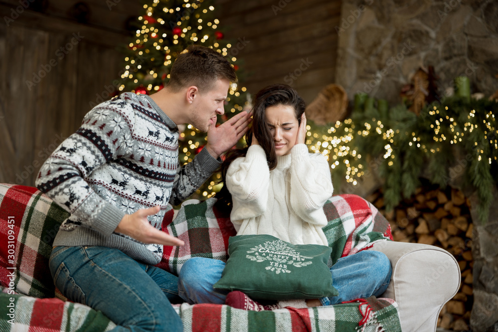 Young couple beautiful woman and handsome man both wearing warm sweaters quarrelling in room decorated for celebrating the new year christmas family drama