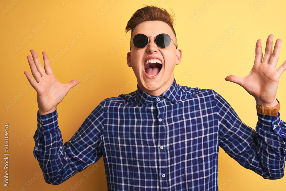 Naklejka premium Young handsome man wearing casual shirt and sunglasses over isolated yellow background celebrating crazy and amazed for success with arms raised and open eyes screaming excited. Winner concept