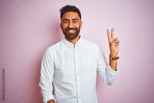Young indian businessman wearing elegant shirt standing over isolated pink background smiling with happy face winking at the camera doing victory sign. Number two.