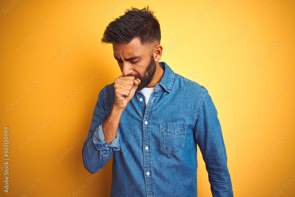 Young indian man wearing denim shirt standing over isolated yellow background feeling unwell and coughing as symptom for cold or bronchitis. Healthcare concept. Young indian man wearing denim shirt standing over isolated yellow background feeling unwell and coughing as symptom for cold or bronchitis. Healthcare concept.