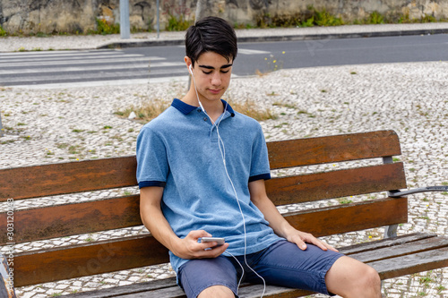 Young boy looks at his smartphone while listening to music. He is sitting on a bench in the city.