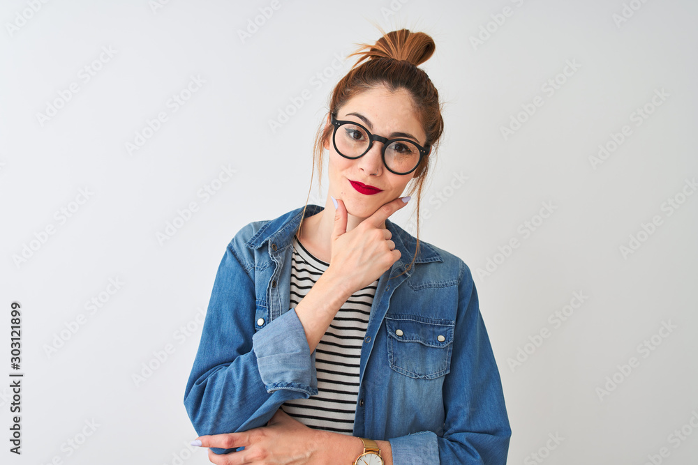 Redhead woman wearing striped t-shirt denim shirt and glasses over isolated white background looking confident at the camera smiling with crossed arms and hand raised on chin. Thinking positive. Redhead woman wearing striped t-shirt denim shirt and glasses over isolated white background looking confident at the camera smiling with crossed arms and hand raised on chin. Thinking positive.