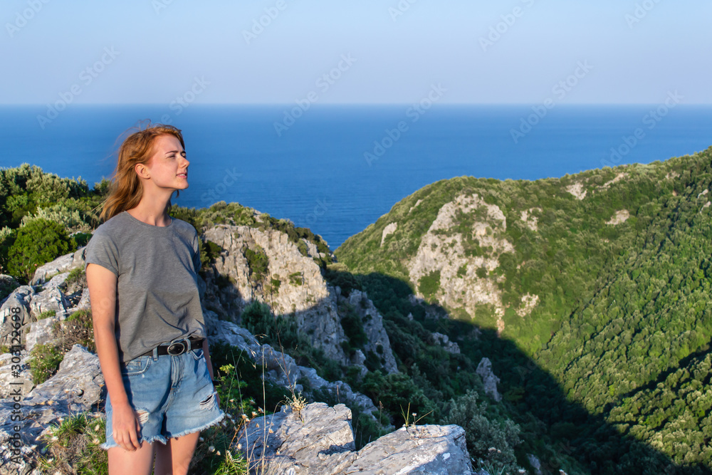 Naklejka premium Red-head young naturally beautiful girl stays in simple summer outfit in relaxation in mountains with blue sea on the back, Greece mountains
